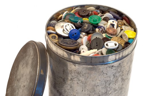 A round metal rubbish bin filled with an assortment of plastic and metal buttons, many of which are various sizes, shapes, and colors including blue, white, black, green, and beige. The bin is positioned outdoors on a paved surface, with a detached metal lid resting against its side. The background is plain white, emphasizing the cluttered contents of the bin, which appear to be collected for disposal or recycling. The scene is neutral and functional, reflecting waste collection activities typical of rubbish removal services, and subtly indicating a focus on small household waste items that [company name] might handle. The bin shows signs of wear with a slightly distressed exterior finish, and the variety of button types suggests they are no longer in use, aligning with the context of clearance or waste management in the Milton Keynes area.