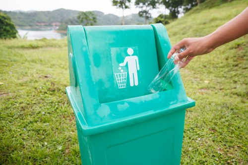 A person’s hand placing a clear plastic bottle into a turquoise bin with a white icon of a person throwing rubbish into a trash can, situated outdoors on a grassy area with a landscape of trees, water, and hills in the background. The bin is designated for waste disposal, possibly part of a waste management initiative in the Milton Keynes area, as seen on the relevant webpage for rubbish removal services by House Clearance Milton Keynes. The environmental setting suggests a park or open space, with the grassy surface extending around the bin and a scenic view beyond, under natural daylight. The image reinforces the importance of proper waste disposal in local outdoor environments, aligning with rubbish removal and recycling efforts common to services in the region.