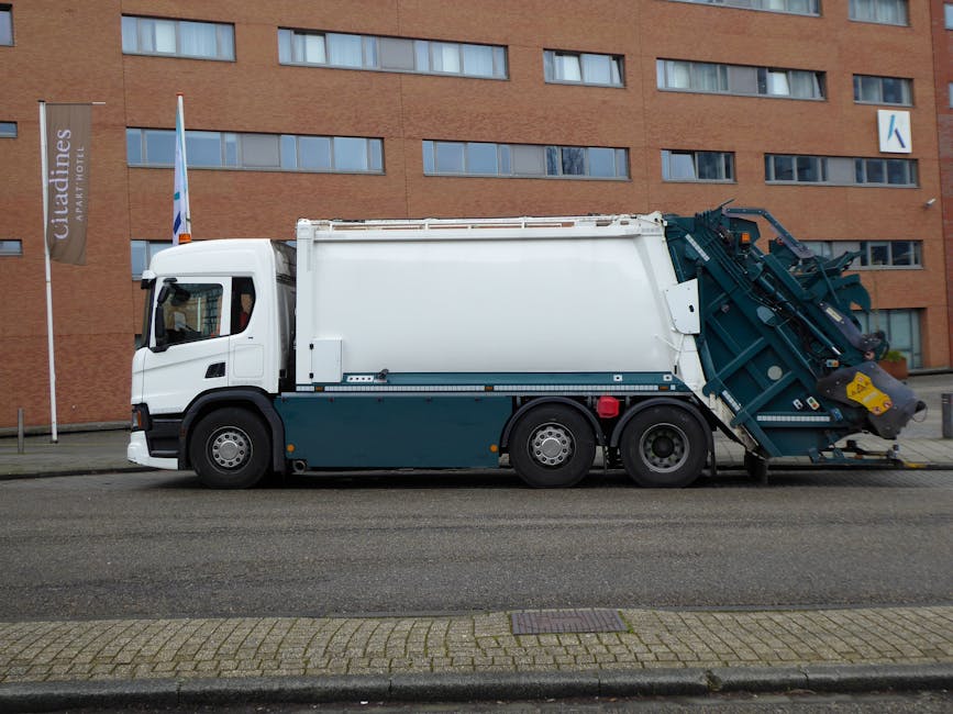 An outdoor urban scene showing multiple open-top rubbish bins and containers overflowing with waste, including cardboard boxes, plastic bags, paper, and packaging materials. The bins are situated on a