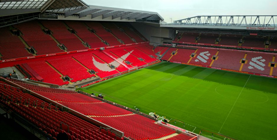 A panoramic view of a large football stadium with a well-maintained grass pitch marked with white lines for play. The seating stands around the field are predominantly red, with some sections featurin